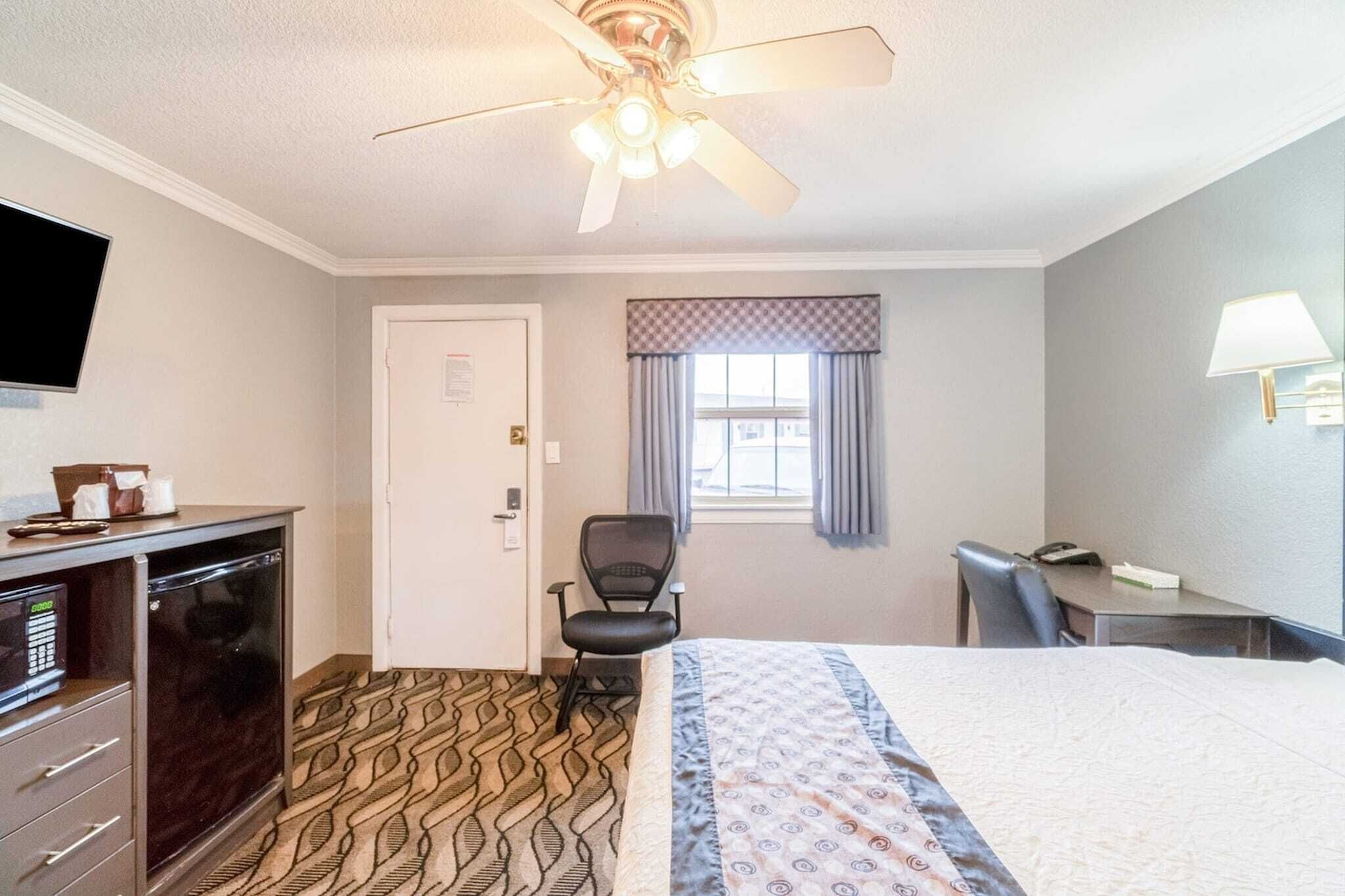 A hotel room with a bed, desk, chair, ceiling fan, TV, and a door.