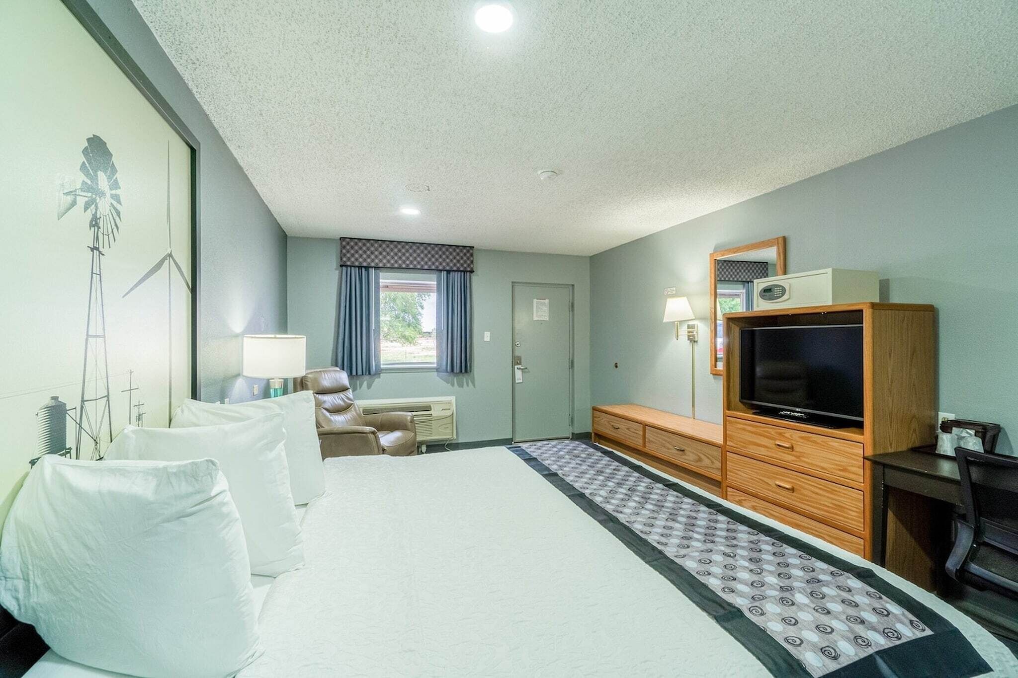 Habitación de hotel con cama, mesita de noche, lámpara, televisión, cómoda y ventana con cortinas.