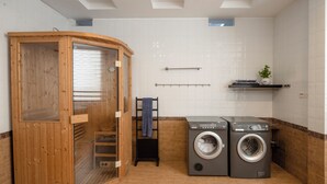 En sauna med trævægge og en glasdør, to frontbetjente vaskemaskiner, et håndklædetørrestativ og en lille hylde med en plante.