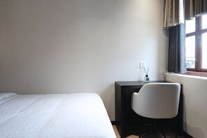 Habitación de hotel con cama, escritorio, silla, ventana con cortinas y toma de corriente.