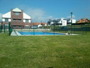 A swimming pool surrounded by a fenced area with a grassy lawn, a playground, and modern buildings in the background. - Cozy one-bedroom apartment with a garden (Miengo)