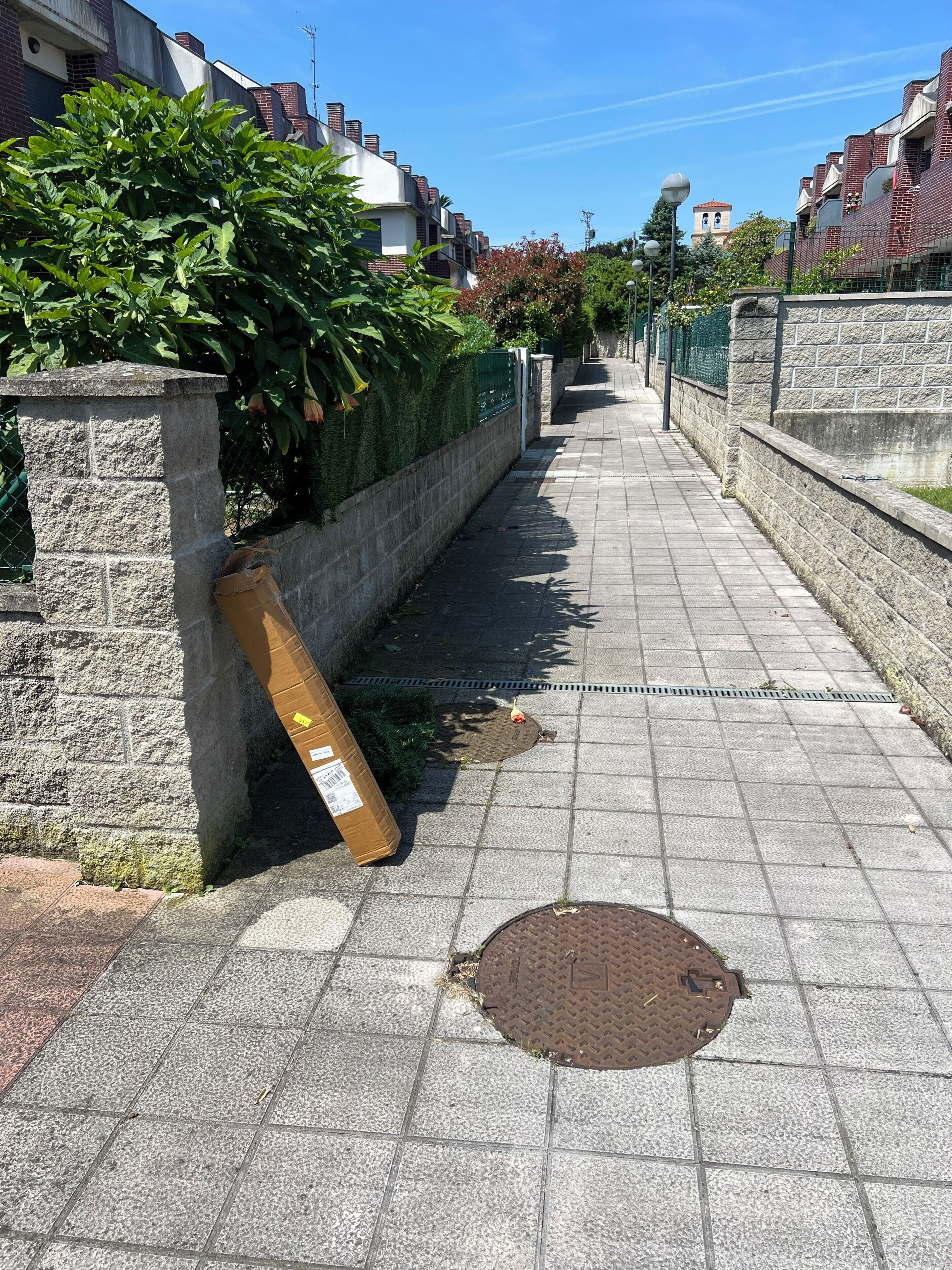 Un trottoir avec un regard d’égout, un mur de pierre et une clôture verte.