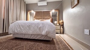 A bedroom with a bed, a nightstand, a mirror, and a wall-mounted air conditioner. - SAOB Property- South AF (Germiston)