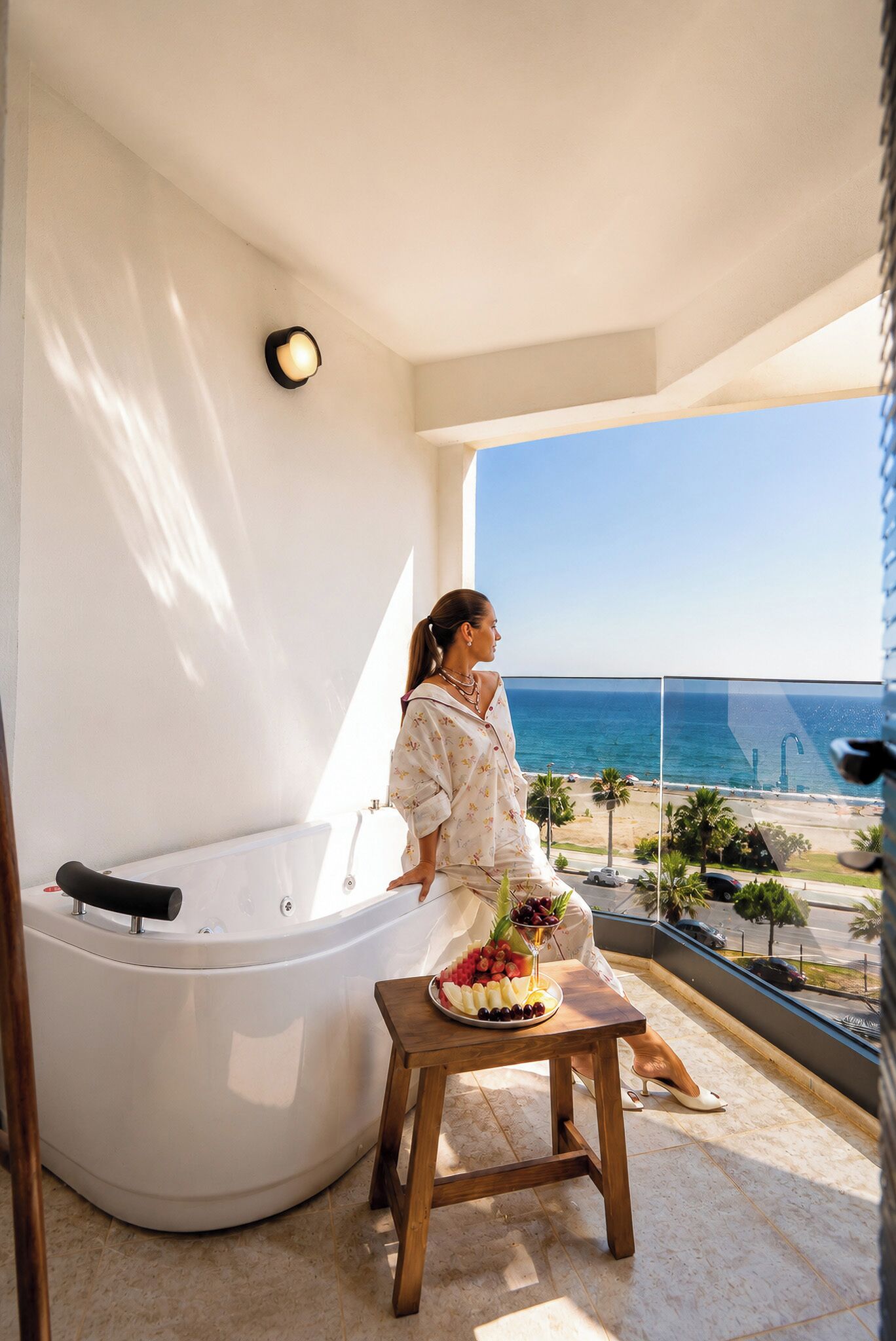 A woman sitting on a balcony with a view of the sea, a hot tub, and a fruit platter on a small table.