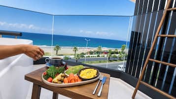 Een balkon met een eettafel, eten op een bord, een kopje, en uitzicht op het strand en de oceaan.
