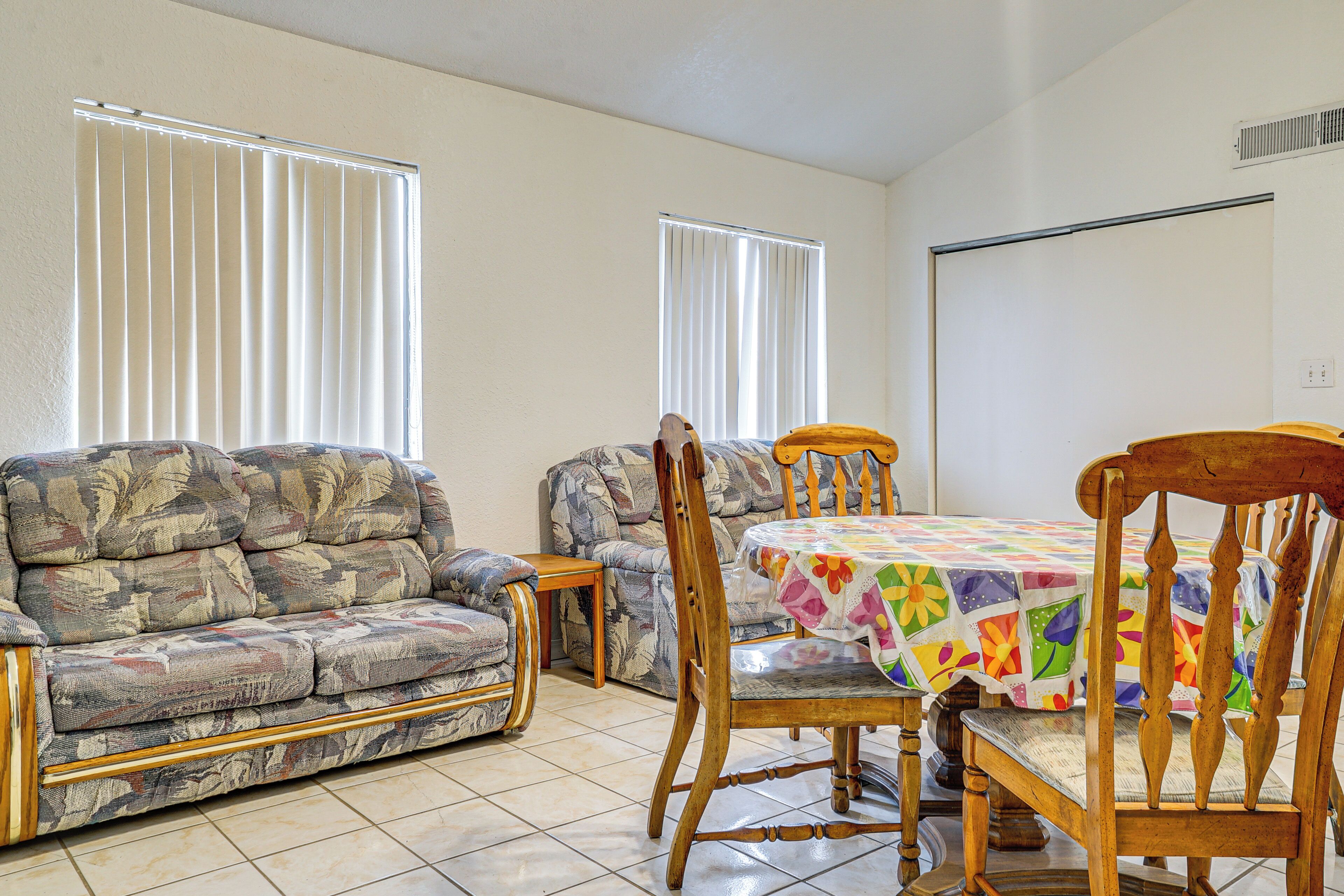 A living room with a sofa, a wooden dining table with a colorful tablecloth, and two chairs.