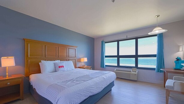 A hotel room with a bed, bedside tables, a desk, a chair, a window with a view, and an air conditioning unit.