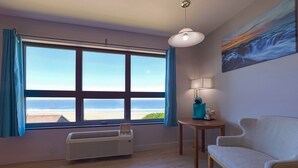 A room with a large window, a sofa, a small table, a lamp, and a painting.