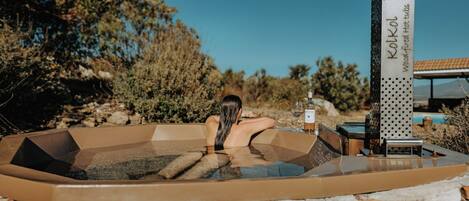 Une femme se détend dans un bain à remous, une bouteille de vin et un verre à la main, entourée d’un mur de pierres et de végétation.