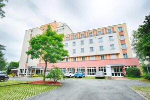 Ein mehrstöckiges Hotel mit zentralem Eingang, einem Parkplatz mit mehreren Autos und einem kleinen Garten mit einem Baum.