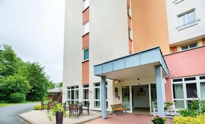 Monikerroksinen hotelli, jossa on katettu sisäänkäynti, ulkona oleva oleskelualue ja hyvin hoidettu puutarha.