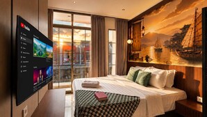 Habitación de hotel con una cama grande, un televisor de pantalla plana en la pared, vistas de la ciudad por la ventana y un mural de un barco a vela en la pared.
