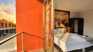 Un cuarto de hotel con balcón, una cama con sábanas blancas, un cuadro de un barco en la pared y vista a la calle.