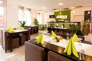 Ravintolassa on useita pöytiä, ja jokaisella pöydällä on vihreät servetit, hopeaesineet ja maljakko täynnä kukkia.