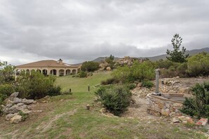 Ein Gebäude mit Kuppel und Bögen, umgeben von einem angelegten Garten mit Kiesweg und kleinem Pool.