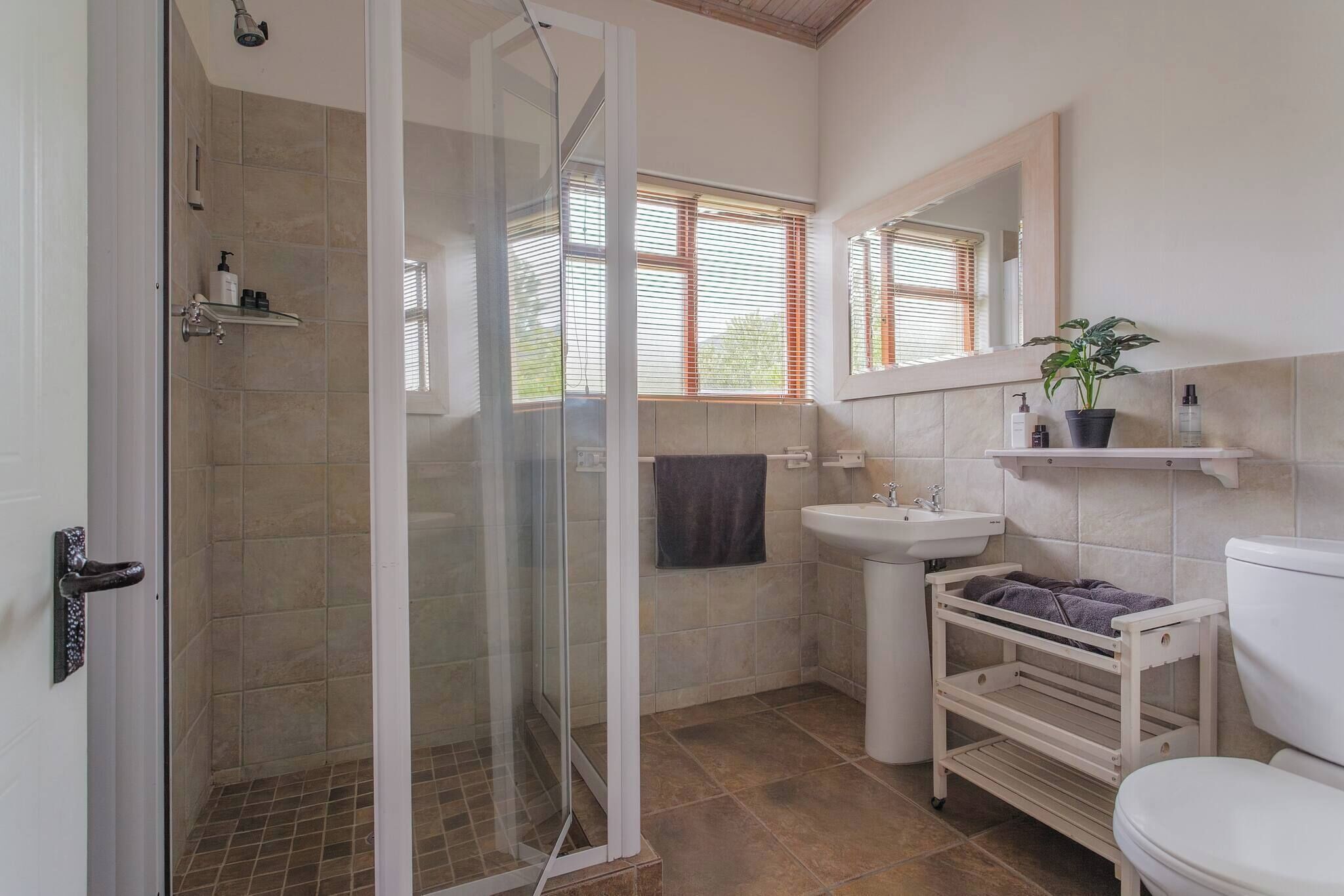 Een badkamer met een douchecabine van glas, een wastafel, een toilet en een raam met zonwering.