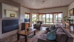 A spacious living room with a large sofa, a coffee table, a television, and a fireplace. - Barton Hammerkop Villa nr Hermanus - HotTub & Pool (Bot River)