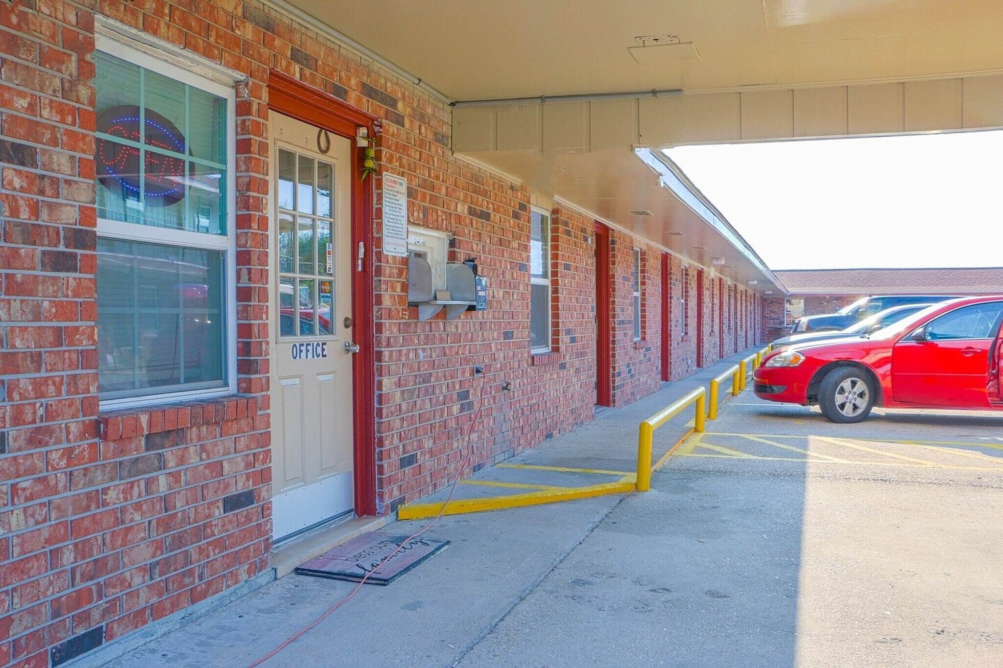 Un edificio de ladrillo con una puerta roja identificada con 'OFFICE', un estacionamiento con pilares amarillos y un auto rojo estacionado allí.