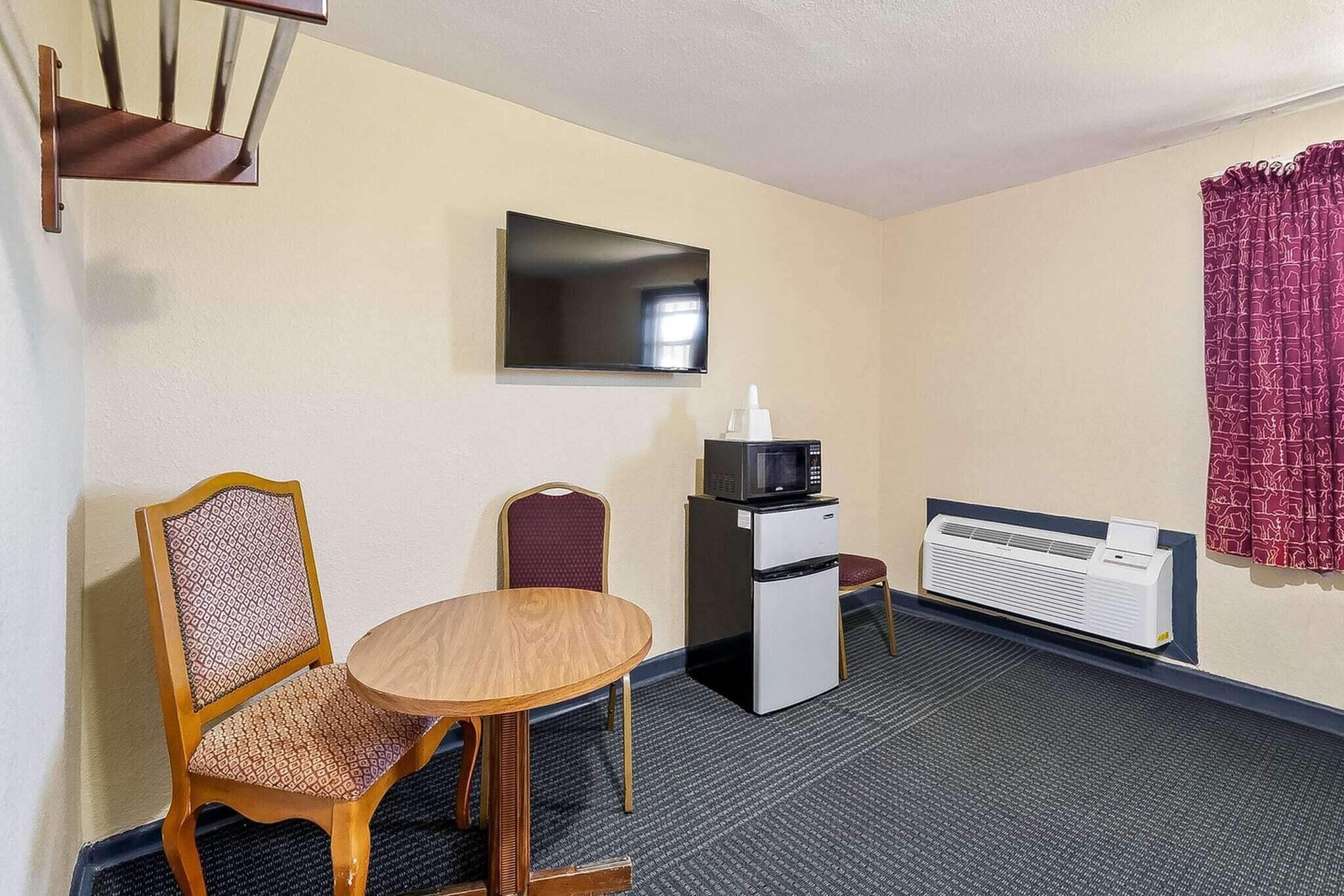 Habitación de hotel con una mesa de comedor, dos sillas, microondas, refrigerador, televisor de pantalla plana, ventana con cortinas y aire acondicionado instalado en la pared.