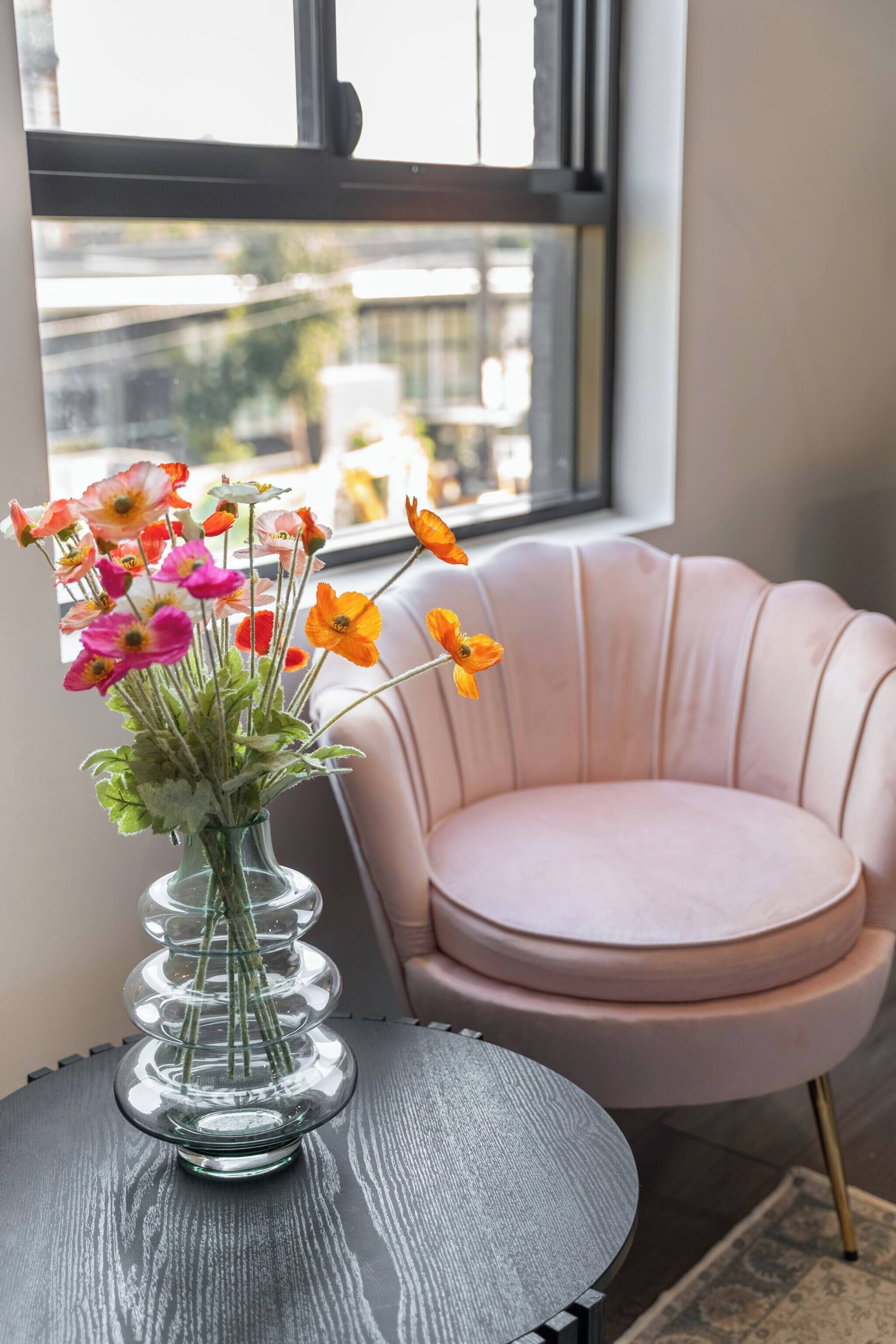A room with a round table, a pink chair, and a vase with flowers.