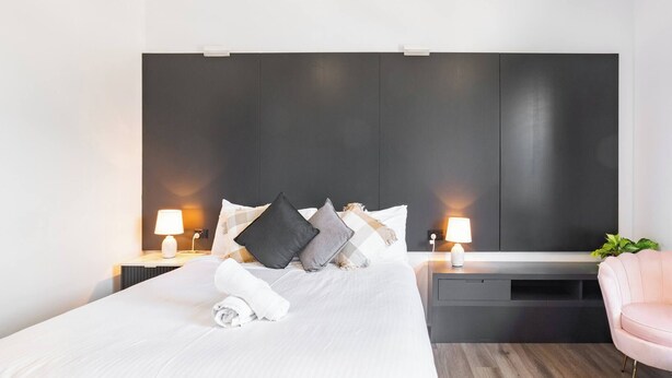 A modern hotel room with a large bed, bedside lamps, a dark headboard, and a pink chair.