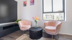 A modern living room with two pink armchairs, a black round table, and a vase with flowers. There is a large window with a city view and two framed pictures on the wall.