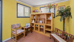 Een kamer met gele wanden, een houten bankje, een klein tafeltje, een boekenplank en een potplant.