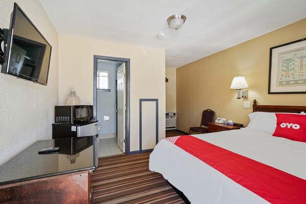 A hotel room with a bed, a desk with a microwave, a television, a door, a nightstand, and a lamp.