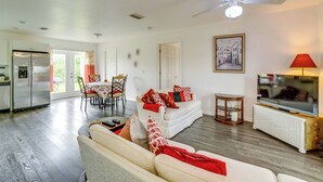 Ein Wohnzimmer mit weißem Sofa, eine Essecke mit Tisch und Stühlen, eine Küche mit Kühlschrank und einem Fernseher auf einem Schrank.