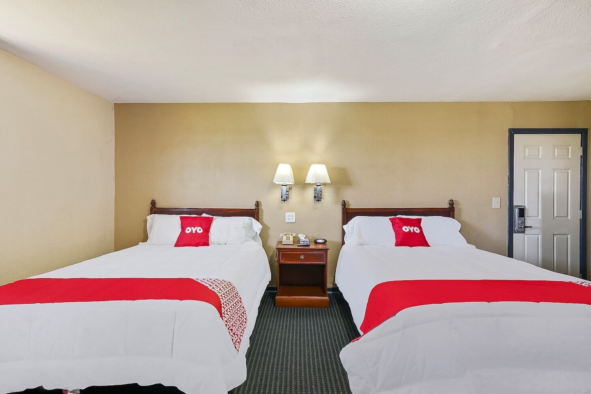 Habitación de hotel con dos camas, cada una con una colcha roja y blanca, un cabecero de madera, una mesita de noche con una lámpara y una puerta a la derecha.