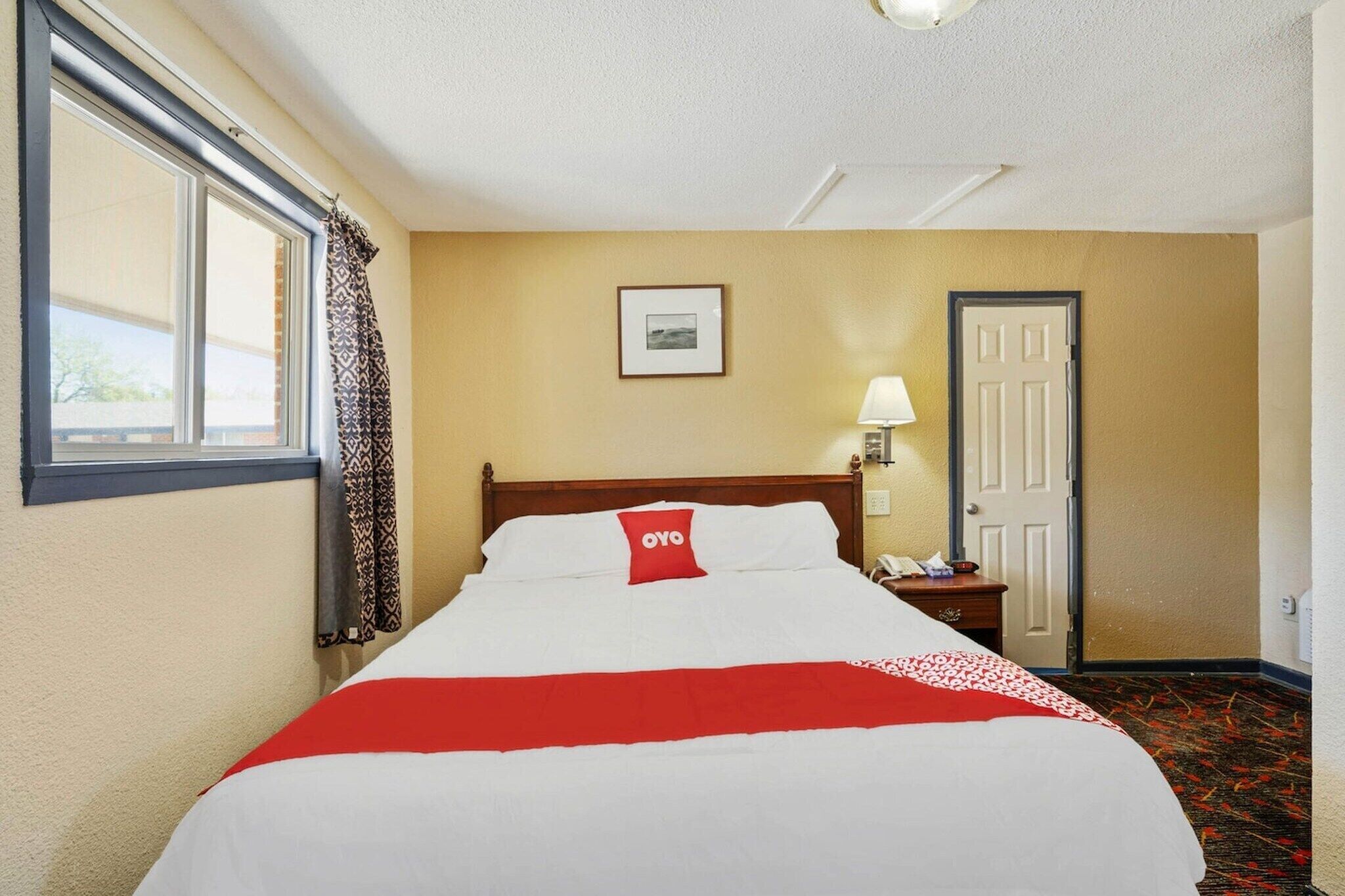 Une chambre d’hôtel avec un lit, une fenêtre avec des rideaux, une porte, une table de chevet avec une lampe et un tableau encadré au mur.