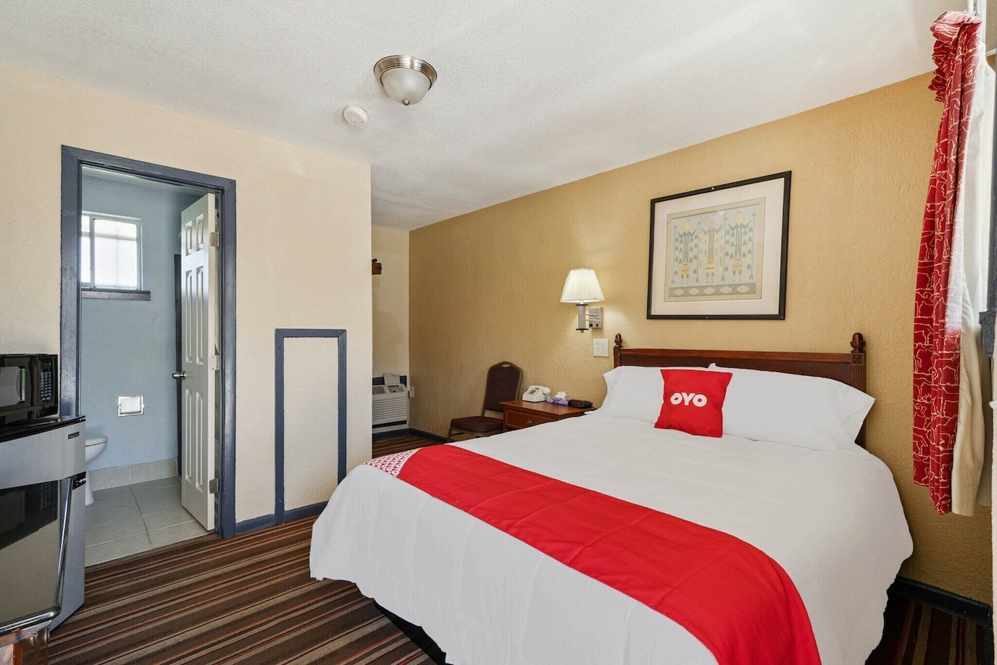 A hotel room with a bed, a desk with a microwave, a bedside lamp, a framed picture on the wall, and a red curtain.
