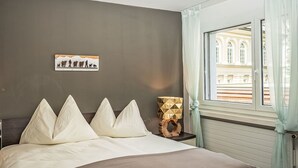 Ein Schlafzimmer mit einem Bett, einem Nachttisch, einer Lampe, einem Bild an der Wand und einem Fenster mit Vorhängen.