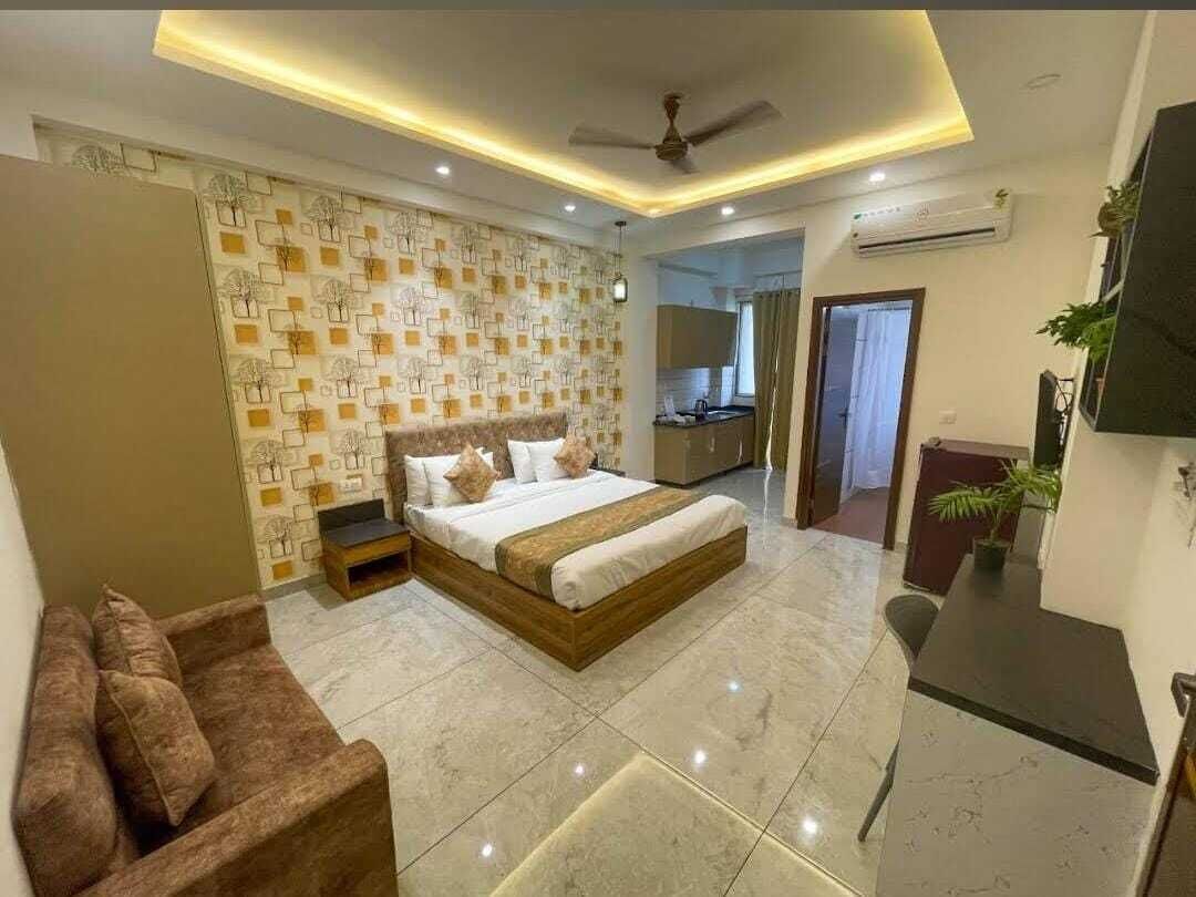A modern bedroom with a large bed, a desk, and a kitchenette in the background.