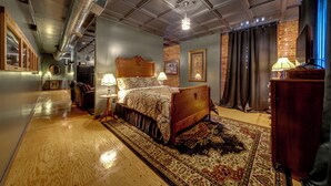 Ein Hotelzimmer mit Holzboden, einem großen Bett mit Kopfteil, einem Nachttisch, einer Lampe, einem Teppich und einer Ziegelwand.