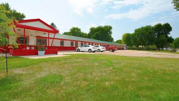 A red and white building with a covered entrance, a parking area with cars, and a grassy lawn.