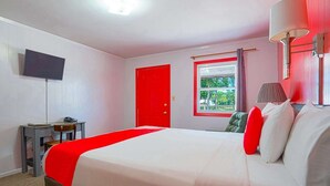 A hotel room with a bed, a desk, a TV, a window with curtains, and a red door.