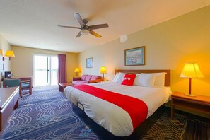 Quarto de hotel com cama, escrivaninha, poltrona, sofá, ventoinha no teto e janela.