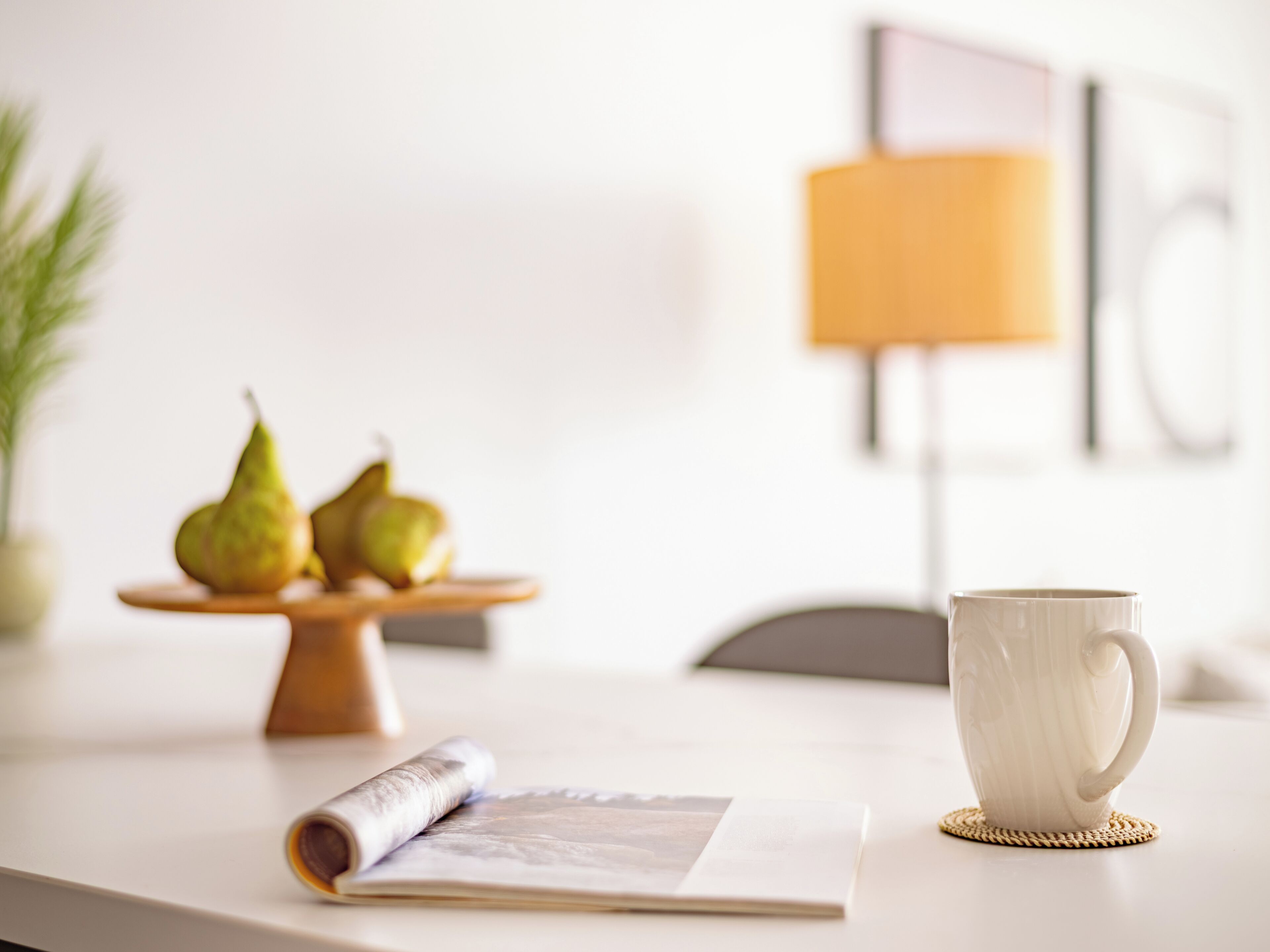 A table with a white mug, a rolled-up magazine, and a wooden fruit stand with pears.
