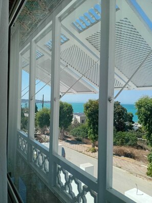 A view through a window with a white balcony railing, a patterned ceiling, and a clear view of the sea and surrounding greenery.