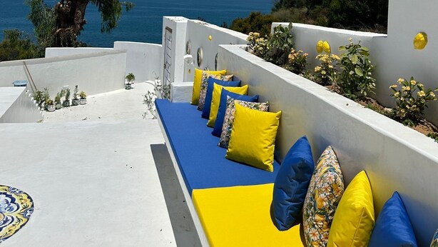 Une terrasse agrémentée d’une rangée de coussins bleus et jaunes, d’un arbre et offrant une vue sur la mer.