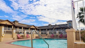 A swimming pool with a metal ladder, surrounded by beige buildings with balconies, and pink chairs. - Premium King Bed Accessible (McAllen TX)