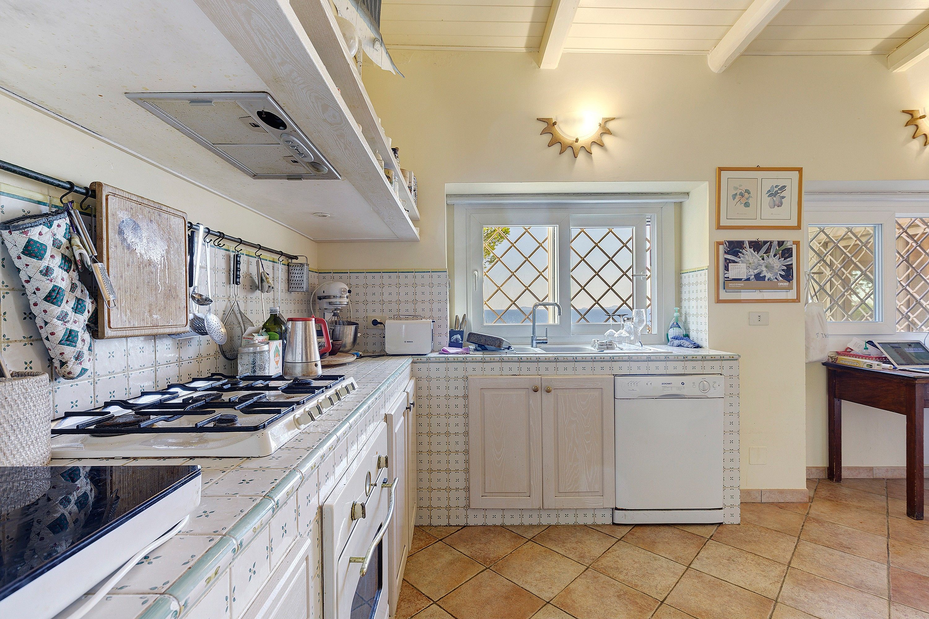 Una cocina con estufa, horno y lavavajillas, una ventana con vistas, y diversos utensilios y electrodomésticos.