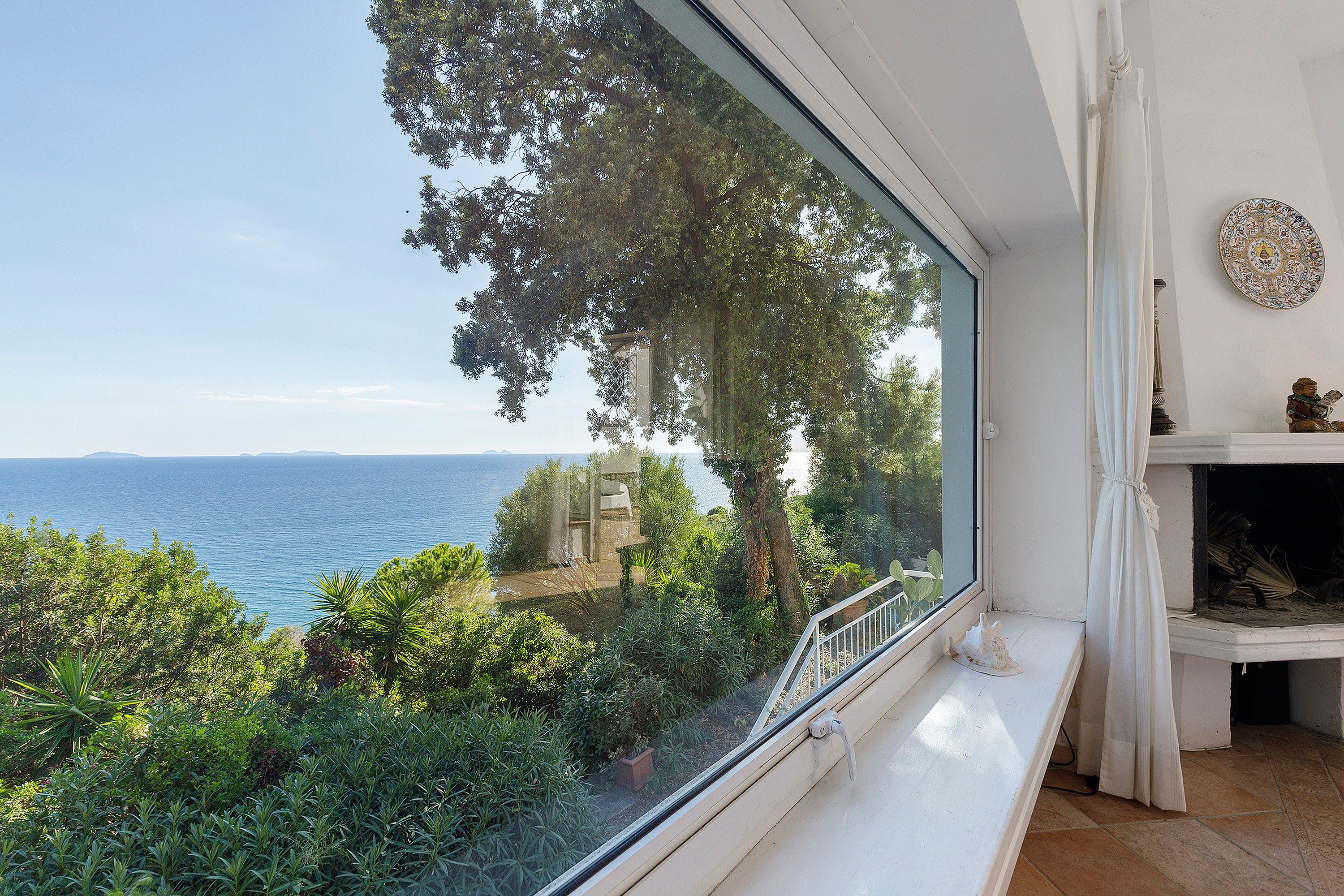 A room with a large window overlooking a coastal landscape, including trees and a view of the sea.