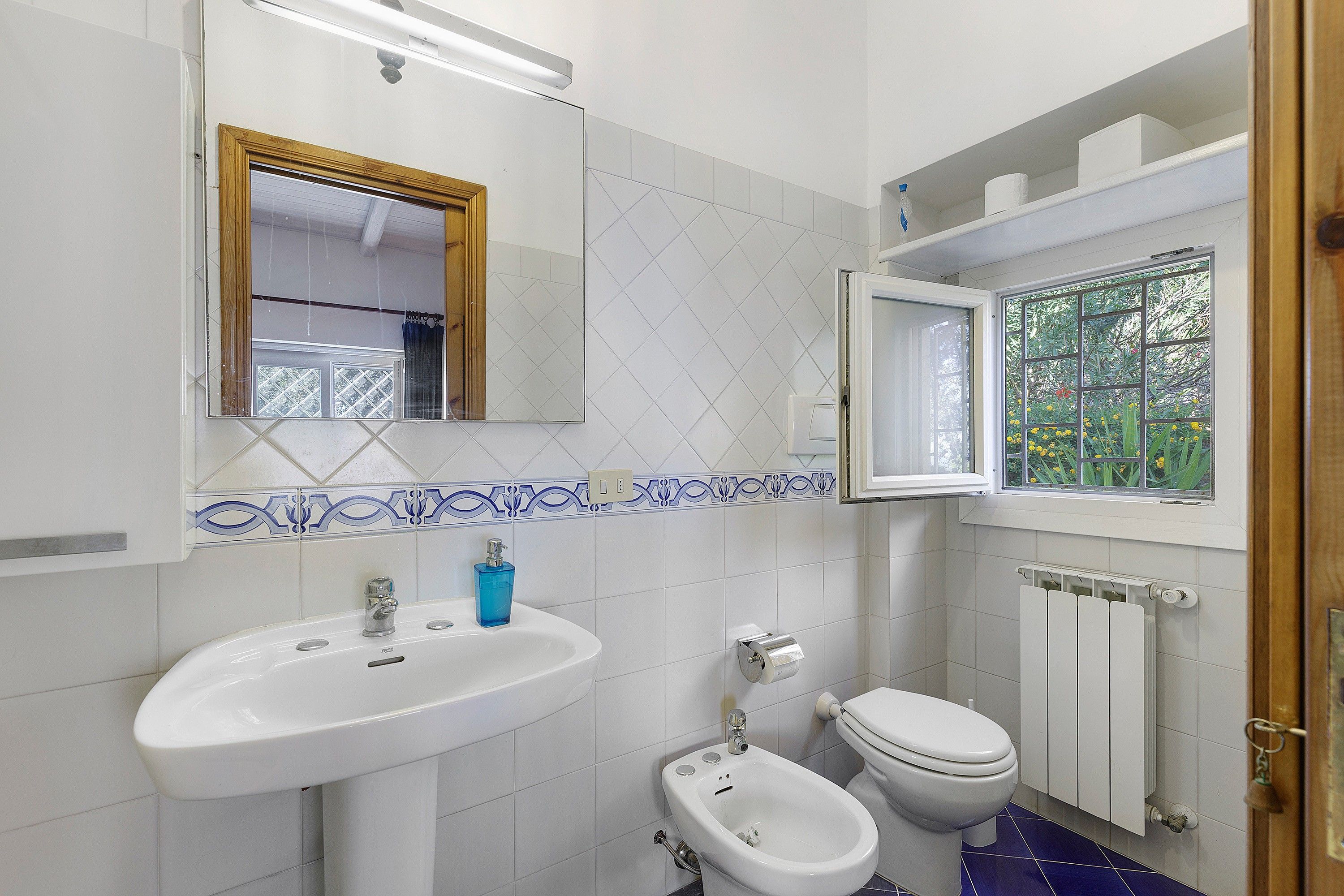 Een badkamer met een wit wastafelmeubel, een toilet, een urinoir en een raam met uitzicht op groen.