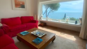 A living room with a red sofa, a wooden coffee table with books, a large window with a view of the sea, and a framed picture on the wall.