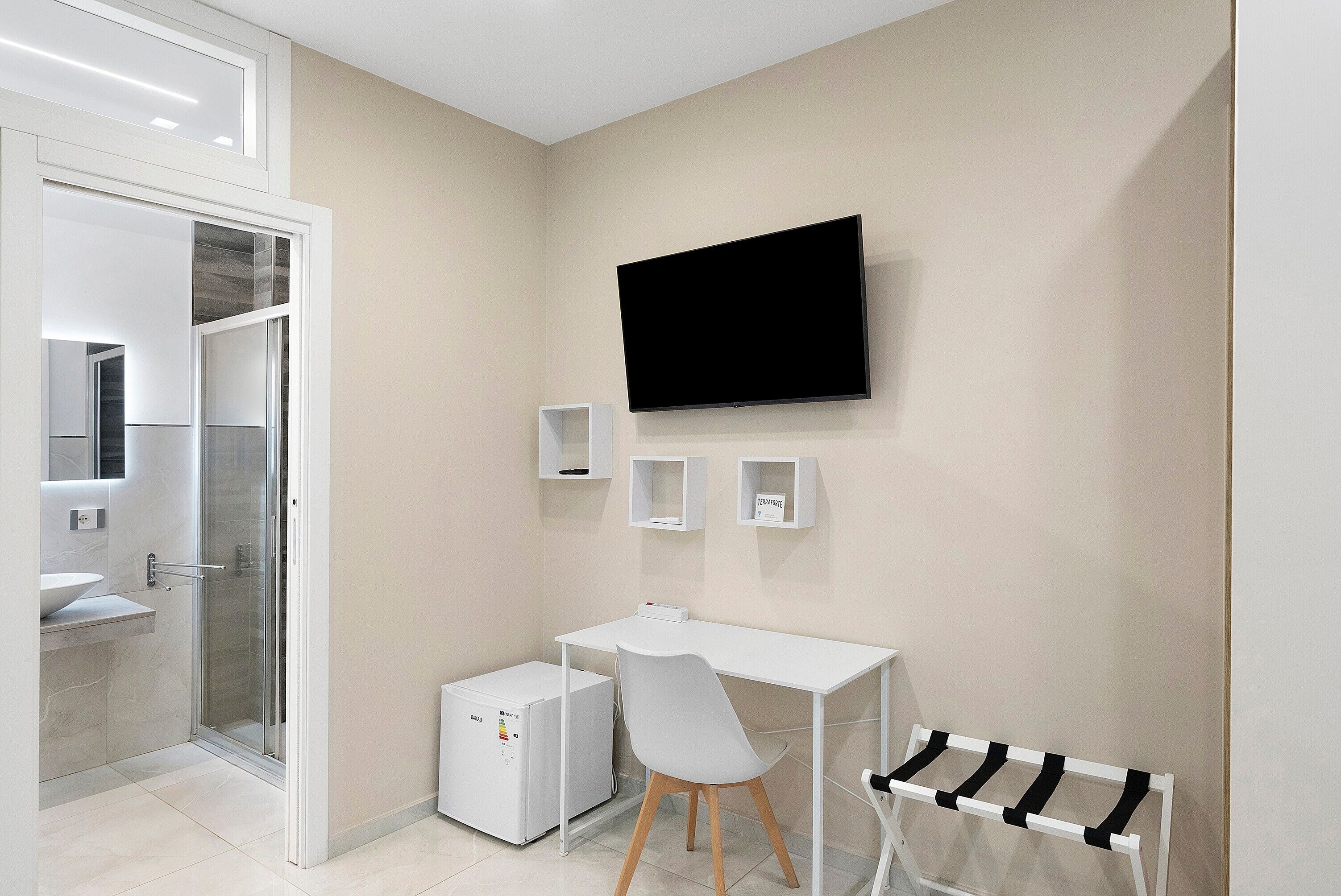 A modern bathroom with a shower, a sink, a toilet, and a white desk with a chair. A flat-screen TV is mounted on the wall.