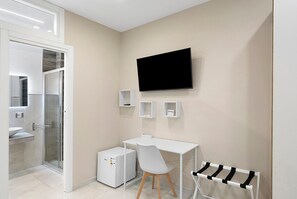 Un baño moderno con ducha, lavamanos, inodoro y un escritorio blanco con silla. Un televisor de pantalla plana está montado en la pared.