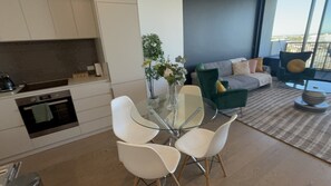 A modern kitchen with white cabinets, a glass dining table with white chairs, and a green sofa in the living area. - Stylish CBD Escape | Sleeps 6 | Pool,Gym & Parking (Adelaide)