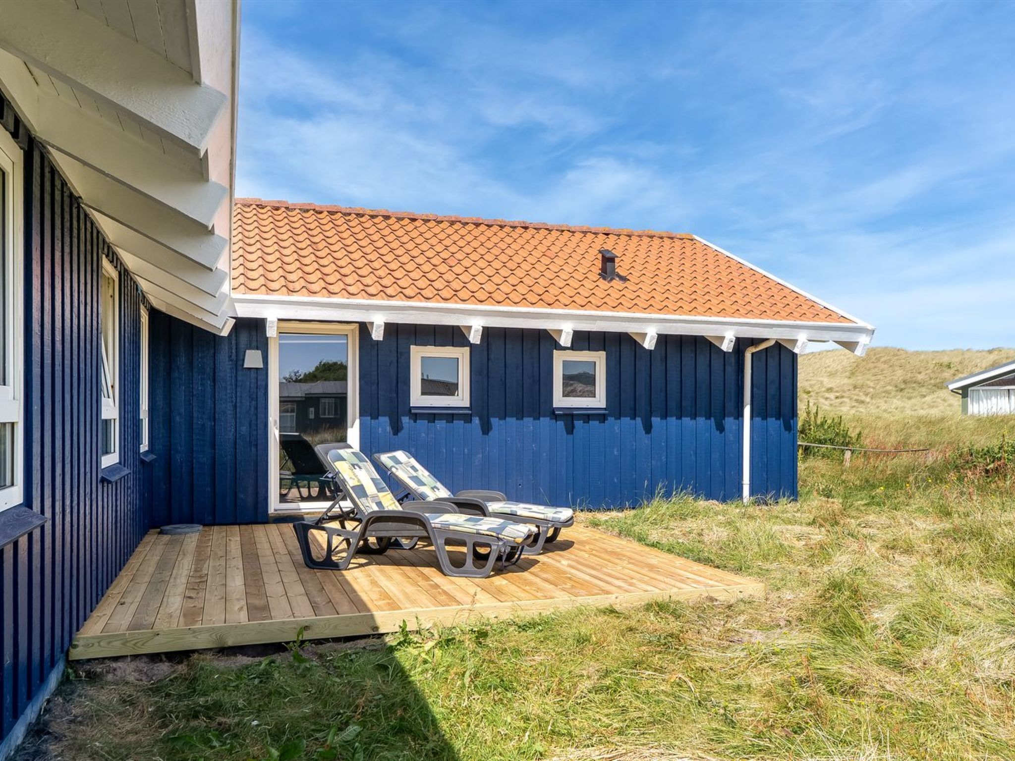 Une maison bleue avec un toit en tuiles, une terrasse en bois et deux chaises longues.